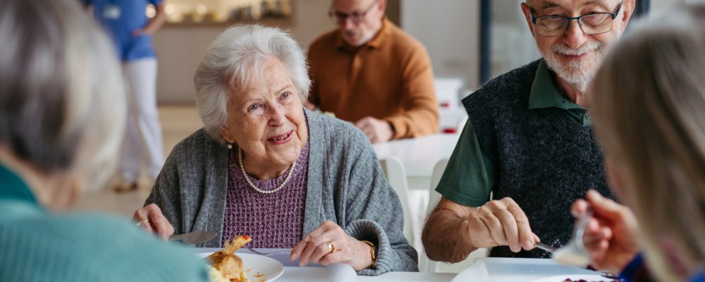 A group of elderly people enjoying a meal together in a bright retirement home dining room with caregiver in background.