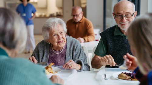 A group of elderly people enjoying a meal together in a bright retirement home dining room with caregiver in background.