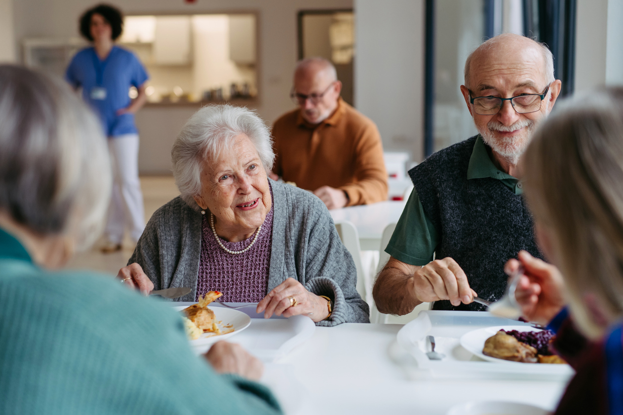 A group of elderly people enjoying a meal together in a bright retirement home dining room with caregiver in background.