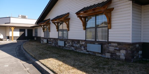 Single-story building with beige siding, stone base, wooden window awnings, driveway and dry lawn
