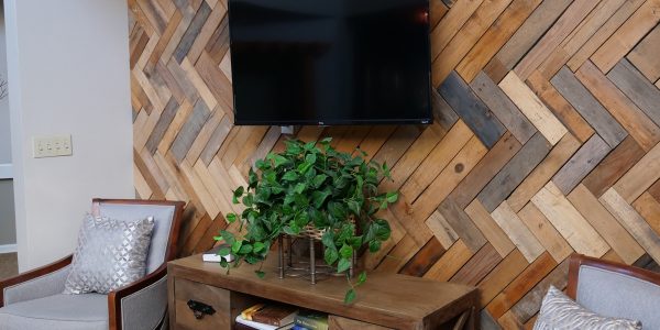 Wall-mounted TV above console table with potted plant and books on herringbone wood accent wall flanked by two armchairs.