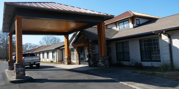 Covered entrance with wooden pillars and stone bases at a low-rise building, pickup truck parked under the porte-cochere.