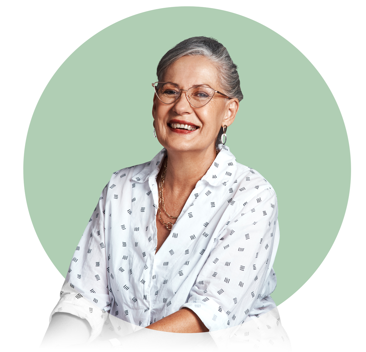 Smiling mature woman in glasses wearing a white patterned blouse and gold necklaces against a muted green backdrop.