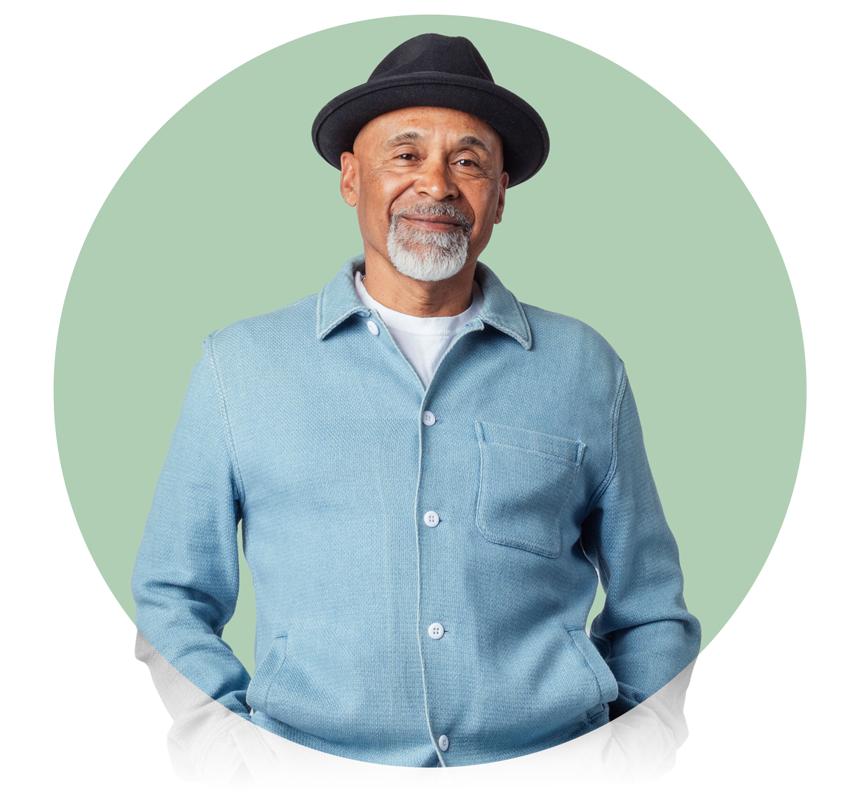 Smiling older man with white beard and black hat wearing light blue jacket, hands in pockets, green circle background