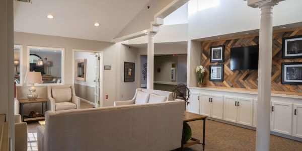Bright neutral-toned lounge with beige sofas, accent wood-paneled wall, TV, columns and soft ambient lighting.