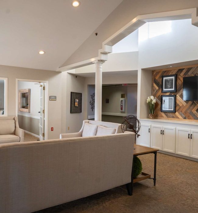 Bright neutral-toned lounge with beige sofas, accent wood-paneled wall, TV, columns and soft ambient lighting.