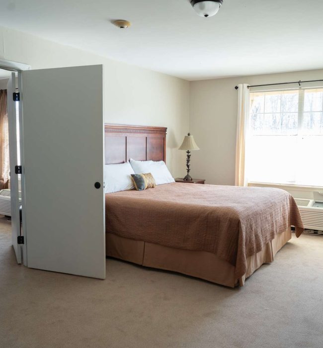 Sunlit bedroom with bed and wooden headboard, bedside lamps, air conditioner under window, open door to adjoining room.