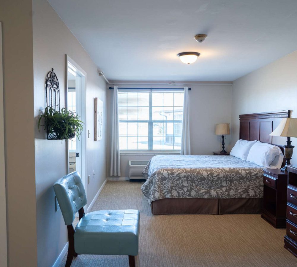 Cozy bedroom with patterned bedspread, wooden headboard, bedside lamps, window with curtains and blue accent chair
