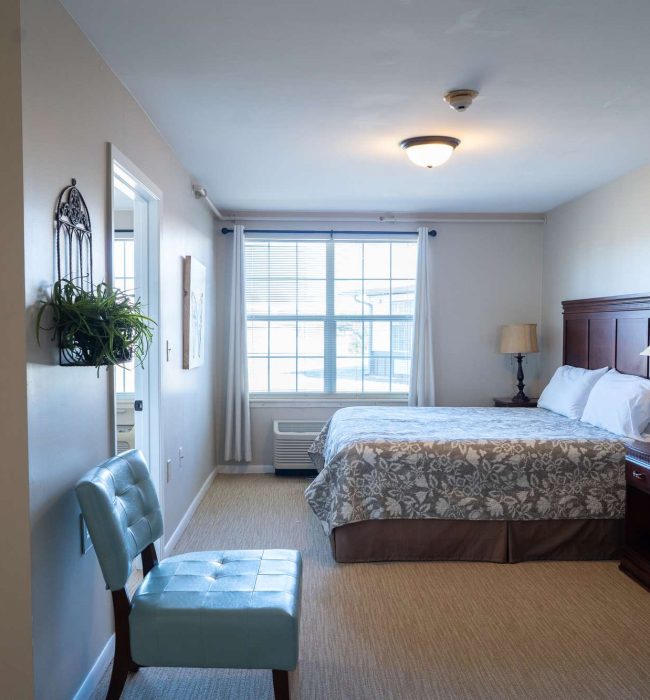 Cozy bedroom with patterned bedspread, wooden headboard, bedside lamps, window with curtains and blue accent chair