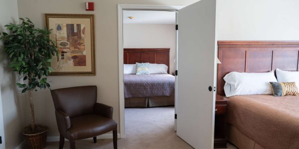 Open doorway between two hotel bedrooms showing brown bedspread, wooden headboard, armchair, potted plant, framed wall art.