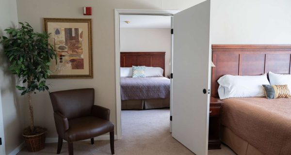 Open doorway between two hotel bedrooms showing brown bedspread, wooden headboard, armchair, potted plant, framed wall art.