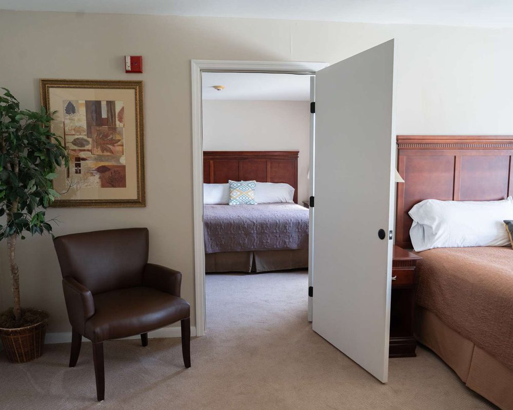 Spacious hotel room with two beds, wooden headboards, open interior door, brown leather chair and potted plant
