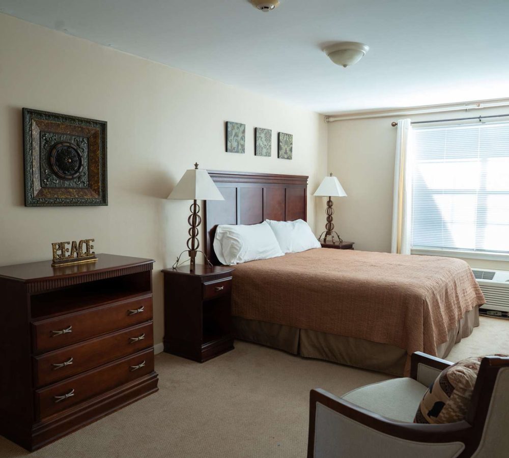 Well-lit hotel-style bedroom with queen bed, wooden headboard, matching nightstands and dresser, window with blinds.