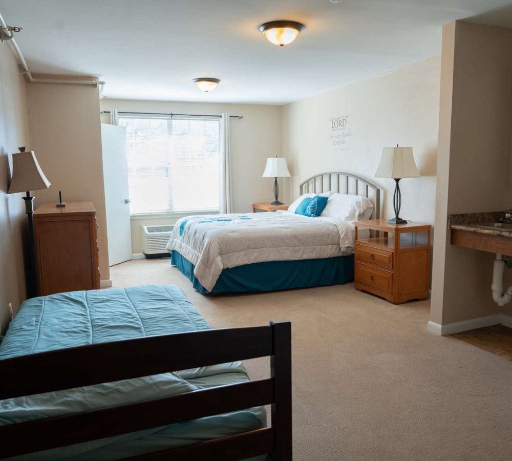 Spacious bedroom with queen and twin beds, wooden nightstands and lamps, large window, beige walls, sink vanity.