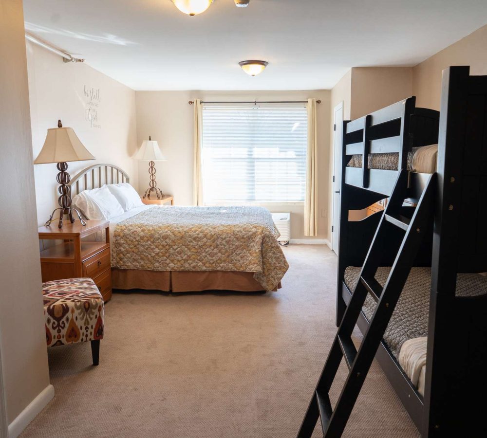 Bright family bedroom with queen bed, dark wooden bunk beds, nightstands and lamps, window with blinds
